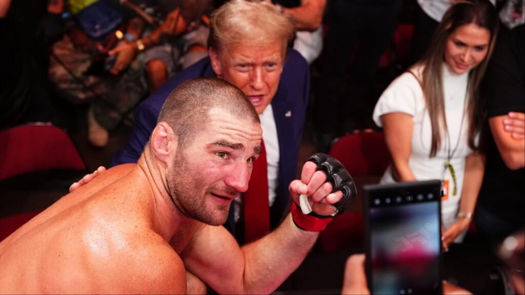 Luchador de MMA sudoroso mira a cámara, Donald Trump se asoma por detrás en evento UFC. Mujer graba con móvil.