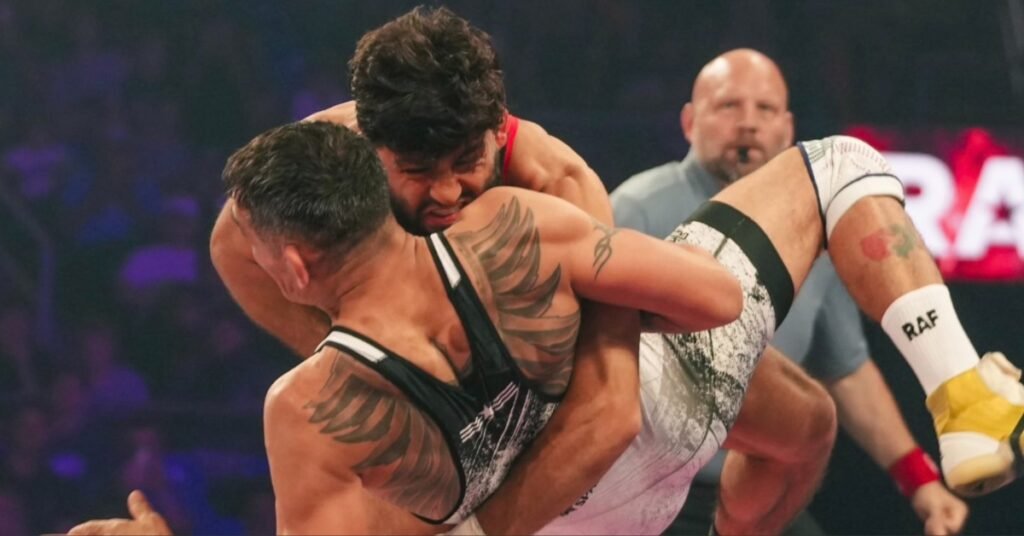 Lucha intensa entre dos atletas. Uno levanta al otro, mostrando tatuajes de alas en la espalda, mientras un árbitro calvo observa el combate.