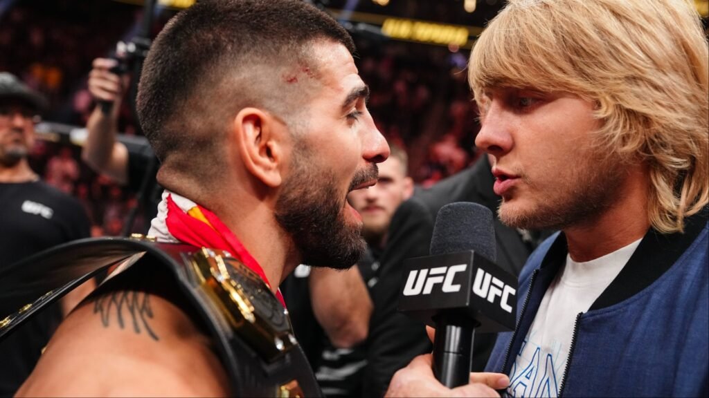 Campeón UFC con cinturón y corte en cabeza, habla intensamente a micrófono. Otro luchador lo sostiene en entrevista poscombate.
