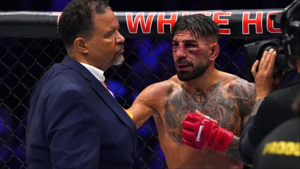 Luchador de MMA con rostro ensangrentado y guantes rojos, dialogando con un árbitro dentro de la jaula.