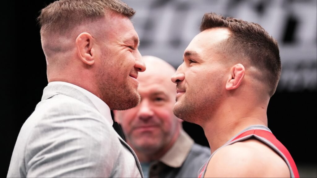 Careo intenso entre dos luchadores de MMA. Uno, de traje gris; el otro, con camiseta deportiva. Dana White observa al fondo.