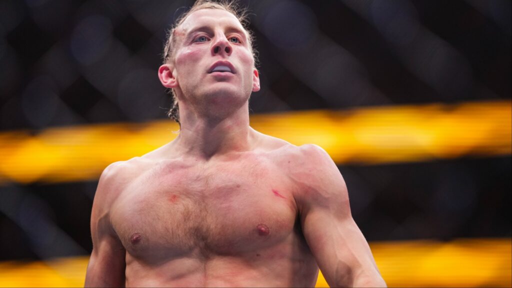 Luchador de MMA, rubio, con torso desnudo y heridas superficiales, mira hacia arriba exhausto en la jaula.