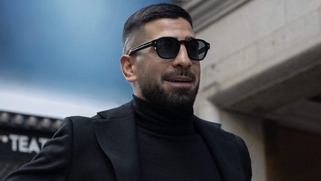 Hombre barbudo elegante con gafas de sol, jersey negro de cuello alto y americana. Sonríe, con fondo azul y un letrero de 'TEAT' borroso.