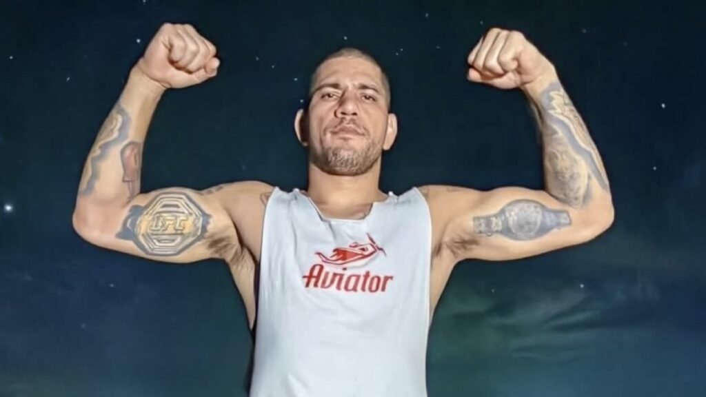Luchador de MMA flexionando bíceps. Lleva tatuajes del logo UFC y un cinturón de campeón. Viste camiseta blanca.