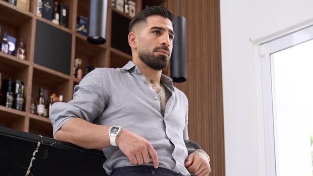 Hombre con barba en camisa gris y reloj blanco, apoyado en mostrador. Fondo con estanterías de bar y ventana.