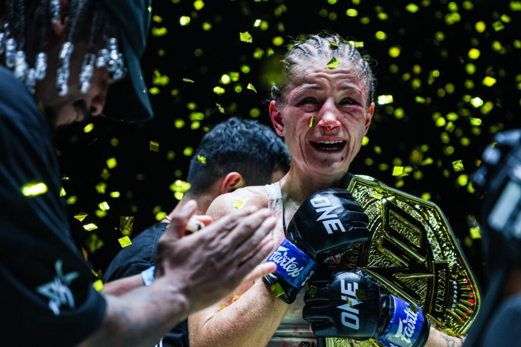 Emocionada campeona de ONE, con guantes y cinturón dorado, lágrimas y sangre en el rostro, celebrando bajo una lluvia de confeti.