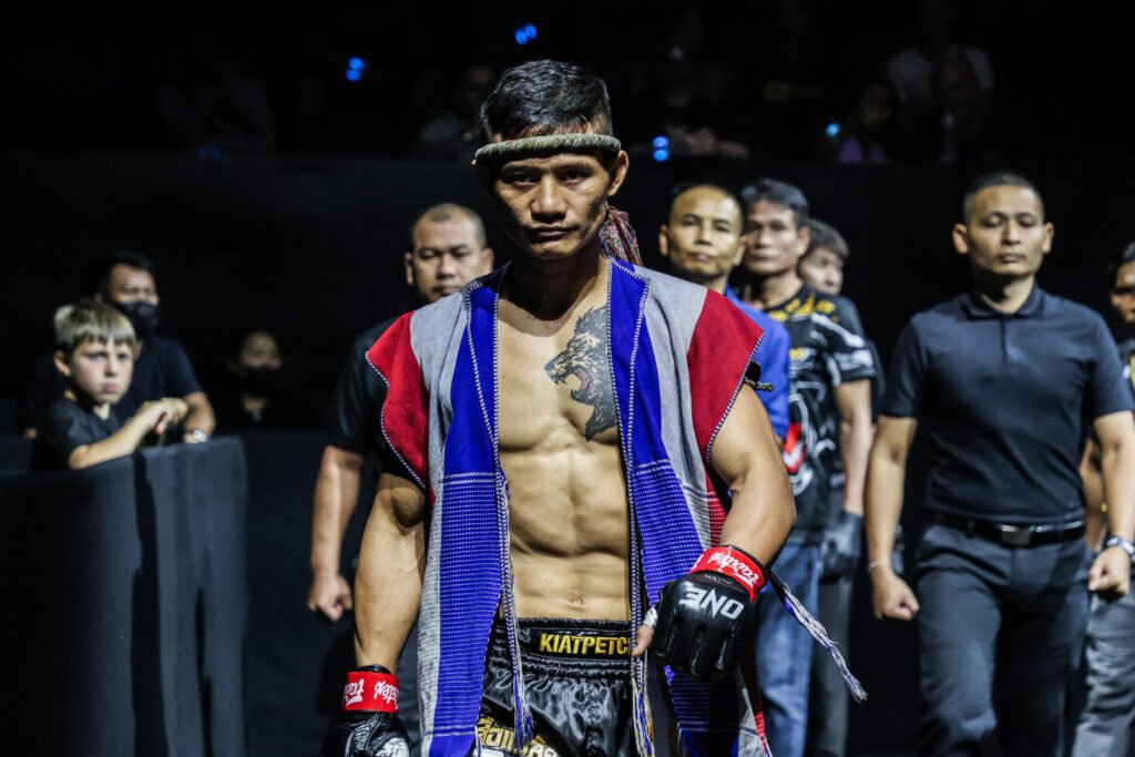Luchador de Muay Thai con mongkol, vestimenta tradicional y guantes MMA, camina al ring con mirada seria.
