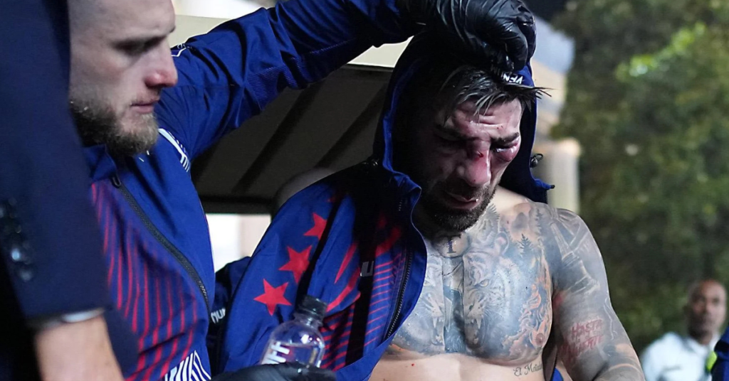 Luchador con la cara ensangrentada, nariz rota y ojos hinchados, siendo asistido tras un combate de MMA. Lleva tatuajes.