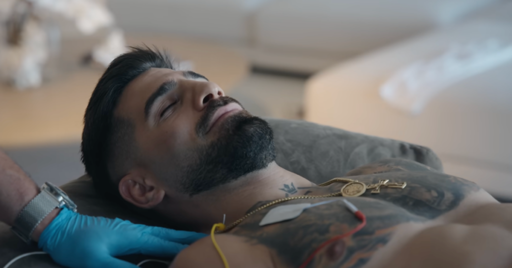 Hombre barbudo y tatuado, ojos cerrados, recostado. Electrodos en el pecho para monitorización médica, una mano con guante azul visible.