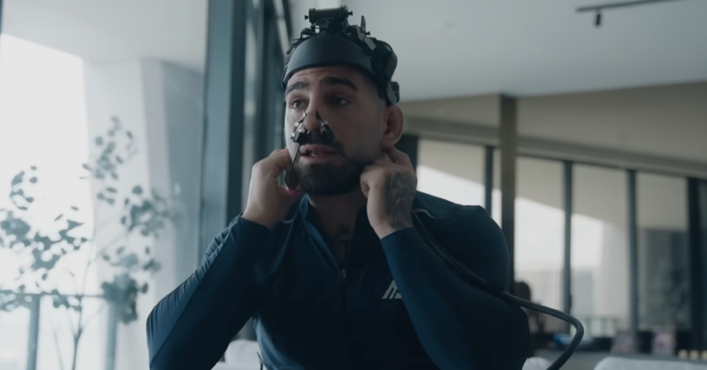 Hombre barbudo con casco de monitorización cerebral y cánula nasal, ajustando sensores en una sala moderna y luminosa.