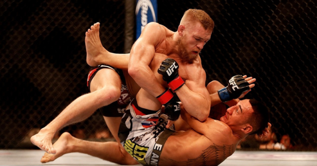 Conor McGregor domina a Max Holloway en el suelo del octógono de UFC durante un combate de MMA.