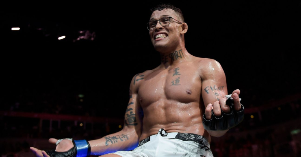 Luchador MMA sudoroso con gafas, tatuajes y guantes sonríe ampliamente en el ring tras una victoria.