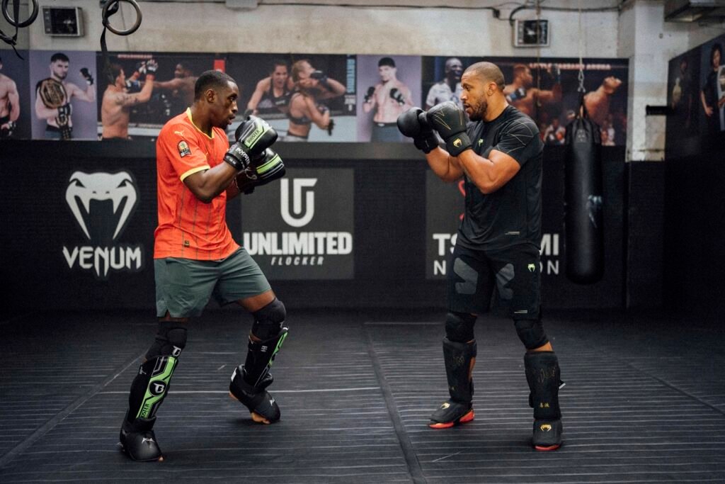 Dos luchadores de MMA en sparring intenso en un gimnasio, equipados con guantes y espinilleras de combate.