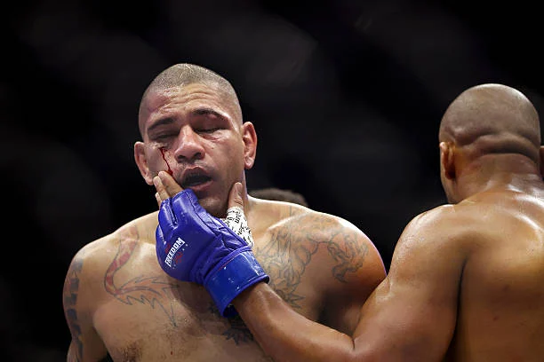 Luchador de MMA con un profundo corte sangrando bajo el ojo izquierdo, tocándose la cara con su guante azul. Otro luchador al lado.
