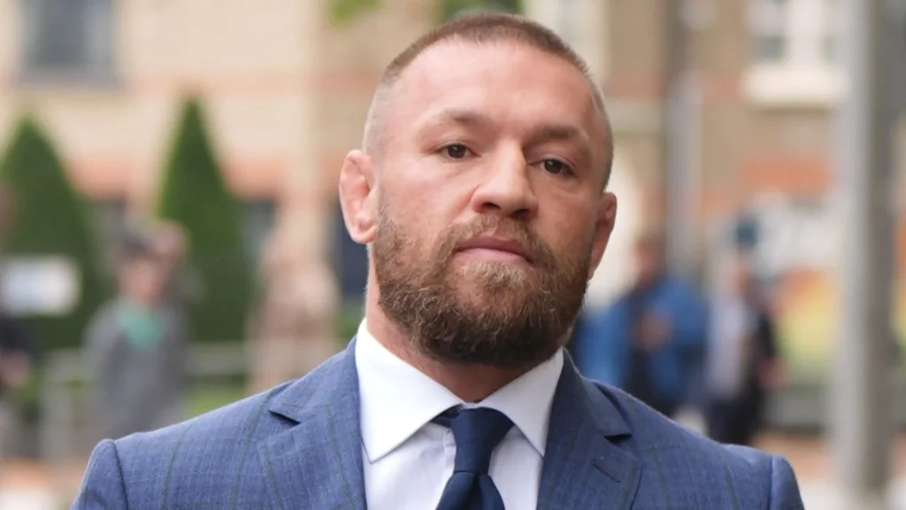 Conor McGregor con barba, traje azul y corbata, mirando directamente a cámara con expresión seria. Fondo urbano difuso.