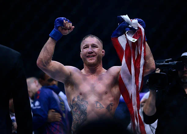 Luchador sonriente de MMA con guante azul alza brazos y bandera estadounidense, celebrando victoria con tatuajes visibles.