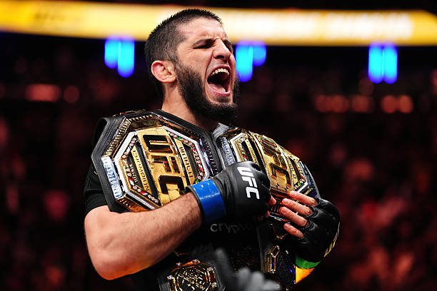 Campeón de UFC celebra eufórico con dos cinturones dorados, gritando con sus guantes de lucha. Arena de fondo.