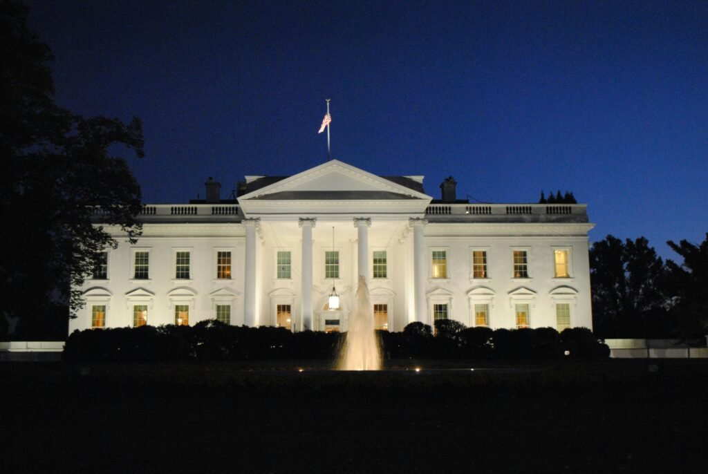La Casa Blanca iluminada de noche, mostrando su icónica fachada, columnas y la bandera de EE. UU. Fuente en primer plano.