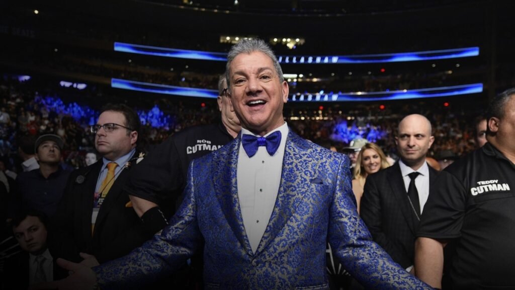 Anunciador Bruce Buffer con elegante traje azul damasco y pajarita, sonriendo en un evento deportivo con público.