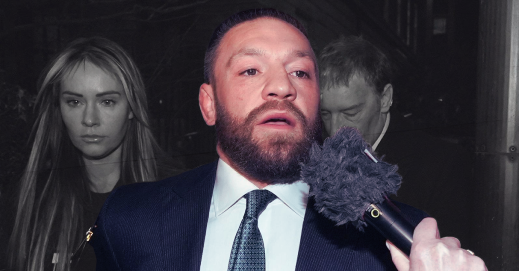 Conor McGregor barbudo, de traje, habla a un micrófono. Mujer rubia seria y un hombre en segundo plano, en tonos apagados.