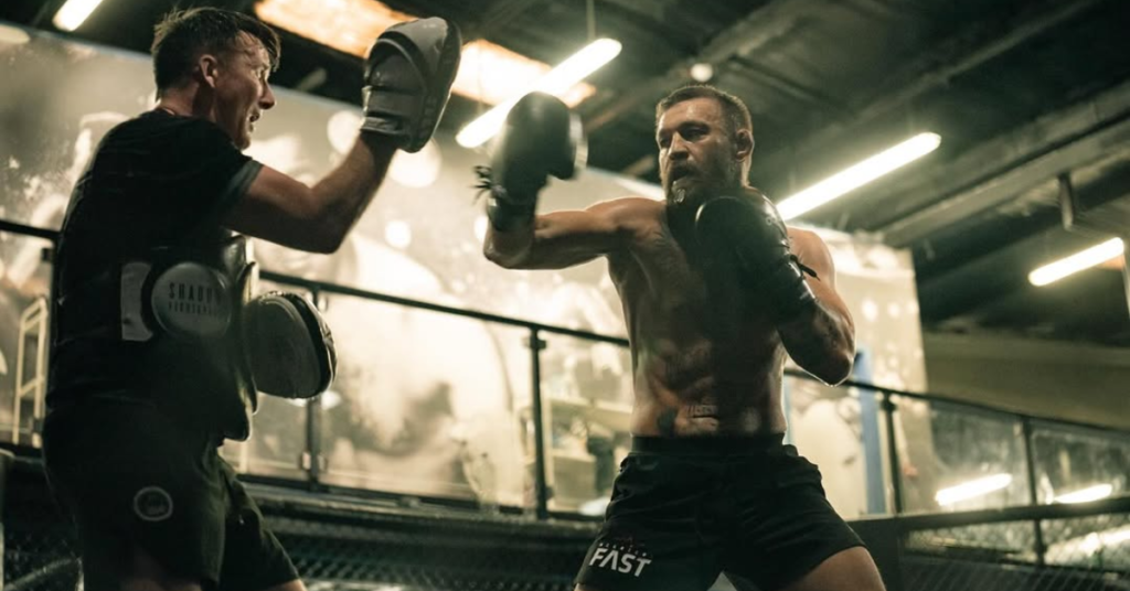 Conor McGregor, sin camiseta, entrena boxeo con su preparador en un gimnasio de MMA, golpeando guanteletas y peto de protección.