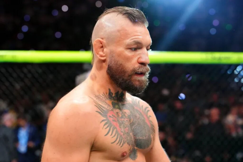 Conor McGregor, luchador de MMA, con barba, mohawk y tatuajes, mira de lado en la jaula de un evento UFC.