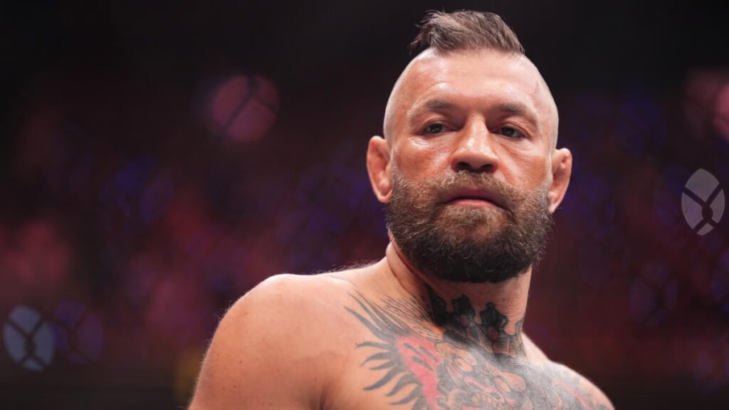 Conor McGregor con barba, corte mohicano y tatuajes en pecho y cuello, mirando fijo. Fondo oscuro de evento UFC.