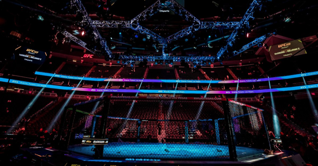 Arena UFC de Las Vegas con el Octágono iluminado en azul y gradas vacías. Pantallas anuncian McGregor vs Holloway 2.