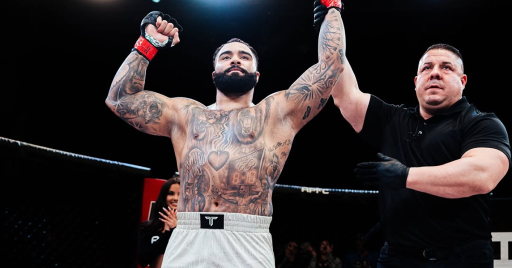 Luchador de MMA tatuado y musculoso con barba celebra su victoria alzando brazos en el ring junto al árbitro.