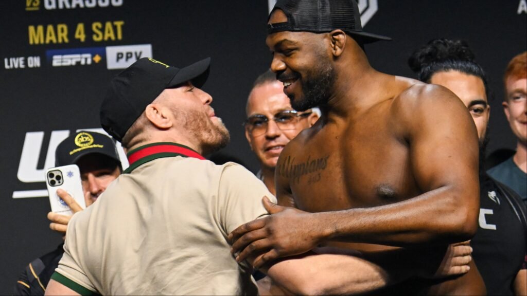 Jon Jones y un rival de UFC cara a cara en un pesaje. Jones sonríe al otro luchador mientras se tocan los hombros.