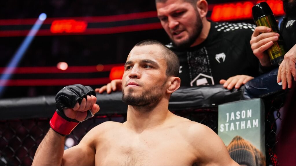 Luchador de MMA con guante Venum negro y puño. Concentrado en la jaula, su entrenador Khabib Nurmagomedov lo asiste.