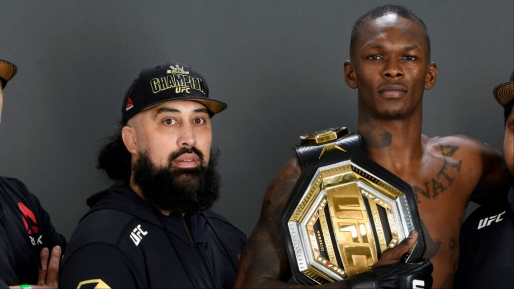 Israel Adesanya, campeón UFC, posa con su cinturón. A su lado, un entrenador con barba y gorra "Champion".