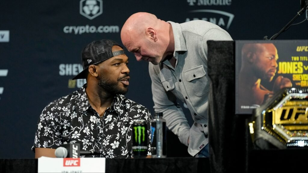 Jon Jones (camisa floral) y Dana White (gris, calvo) conversan en una rueda de prensa de UFC, con logos y cinturón de campeón.