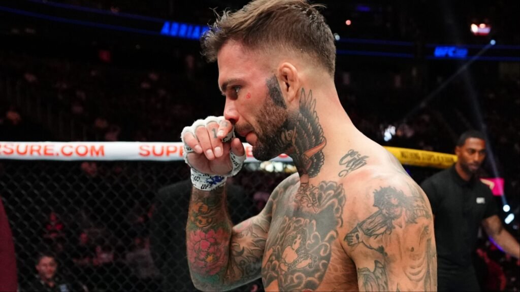 Luchador tatuado de MMA con guantes y barba, pensativo en el octágono de la UFC. Se ven tatuajes en cuello y brazos y marcas rojas bajo el ojo.