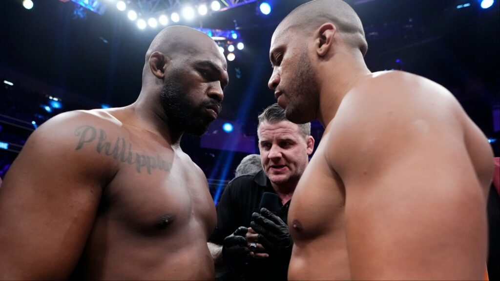 Intenso careo entre dos fornidos luchadores de MMA, mirándose fijamente, con un árbitro interviniendo. Ambiente de combate.