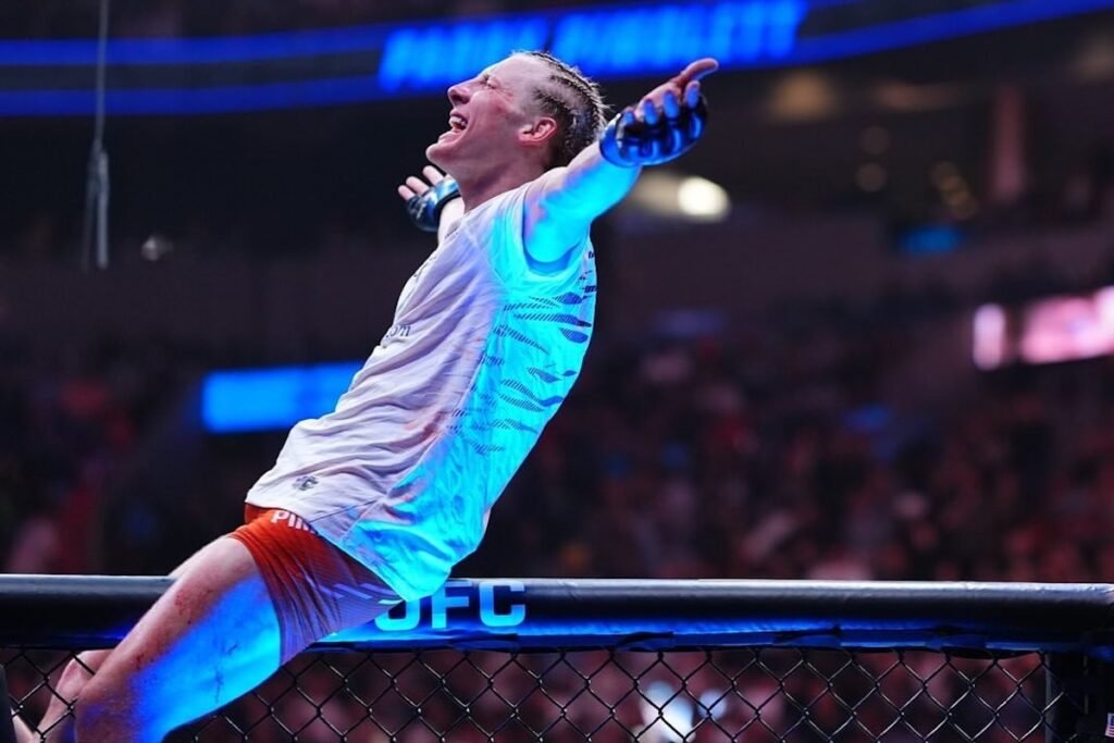 Paddy Pimblett, luchador de UFC, celebra eufórico su victoria con brazos abiertos en el octágono, bañado en luz azul.