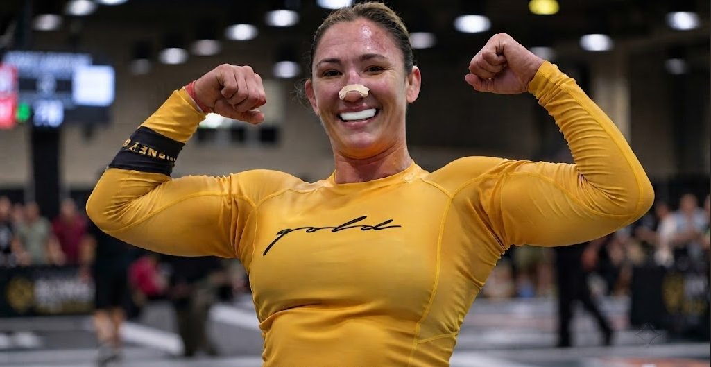 Paige Clymer, atleta de combate, sonríe y flexiona bíceps, con camiseta amarilla 'gold', protector bucal y vendaje nasal.