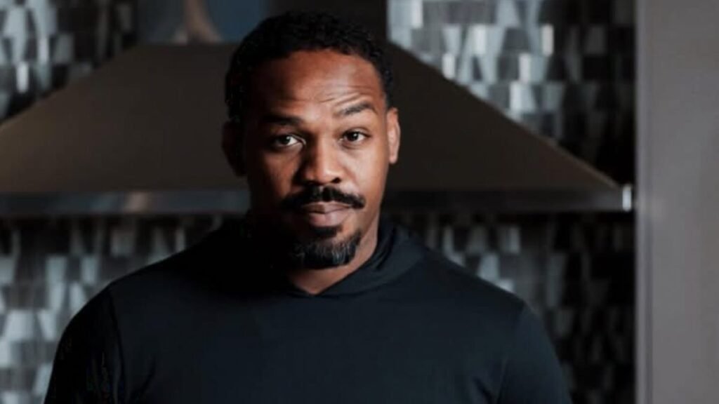 Jon Jones, luchador, con sudadera negra y perilla, mirando serio a cámara. Cocina moderna con campana extractora y azulejos.