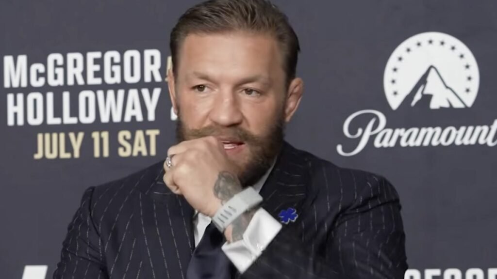 Conor McGregor, barbudo, en un evento de lucha el 11 de julio. Viste traje, con "McGregor Holloway" y Paramount de fondo.