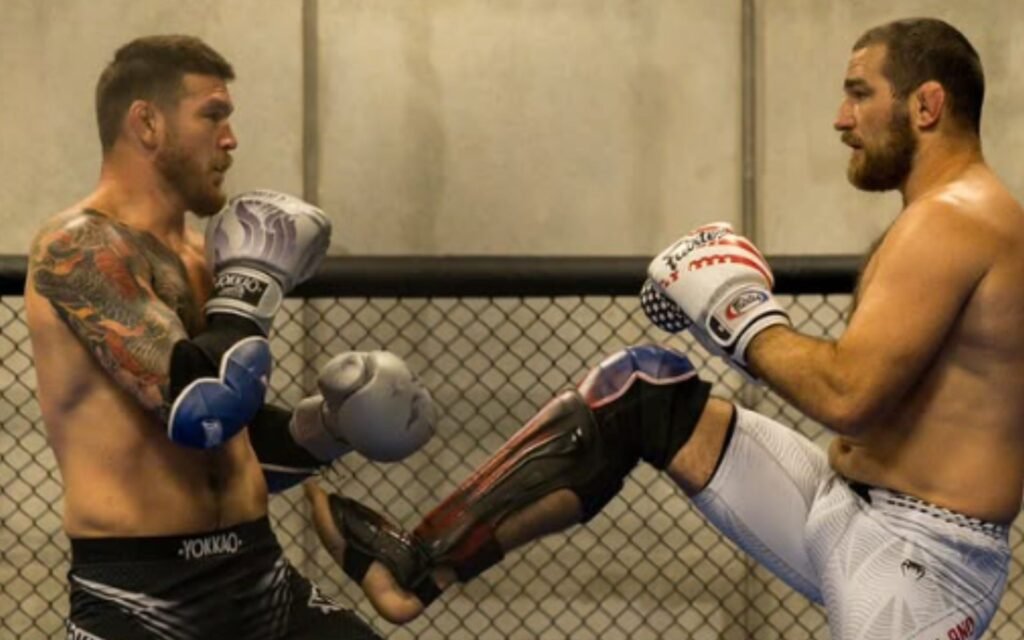 Dos luchadores de MMA con guantes y espinilleras intercambiando patadas bajas en jaula. Entrenamiento de kickboxing con Sean Strickland.