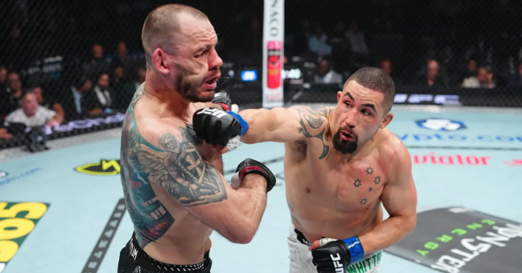 Combate UFC: Luchador tatuado y barbudo impacta con un puñetazo en el rostro de su oponente, con salpicaduras y público borroso en el octágono.