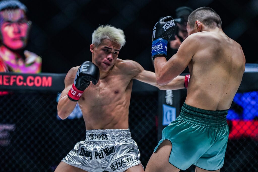 Combate de MMA: Zhang Pei Mian (pelo rubio, shorts plateados) en plena acción, golpeando a su oponente de espaldas con shorts turquesa.