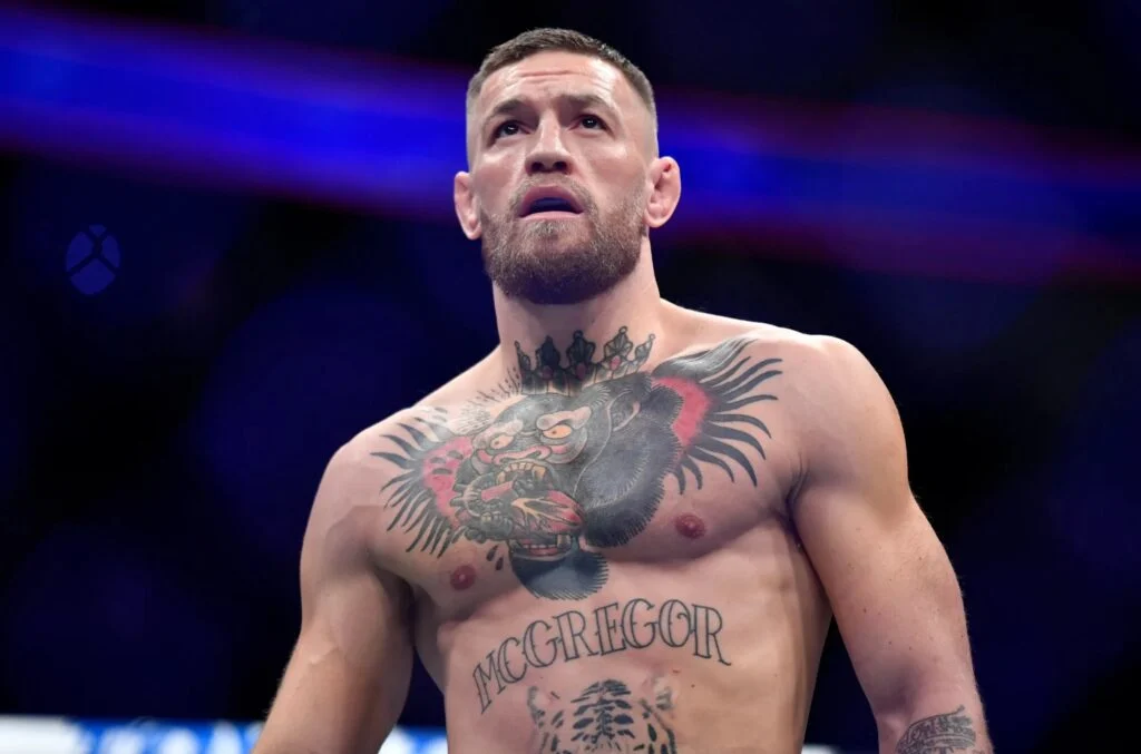 Conor McGregor, con barba, sin camiseta y tatuajes de gorila coronado y su nombre, mira arriba en una arena de MMA.
