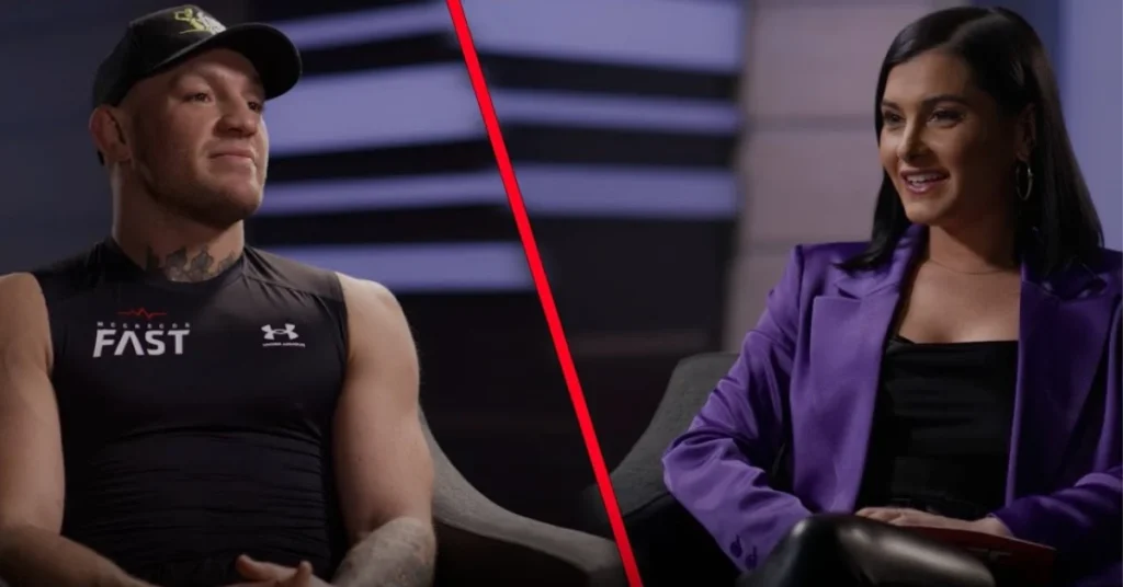 Conor McGregor con camiseta Under Armour "FAST" y gorra, entrevistado por una mujer sonriente con blazer morado.