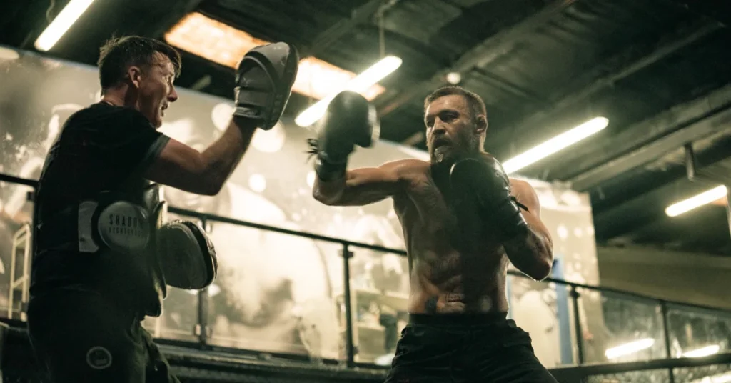 Conor McGregor, sin camiseta y con guantes de boxeo, lanza un puñetazo al peto de su entrenador en un gimnasio.