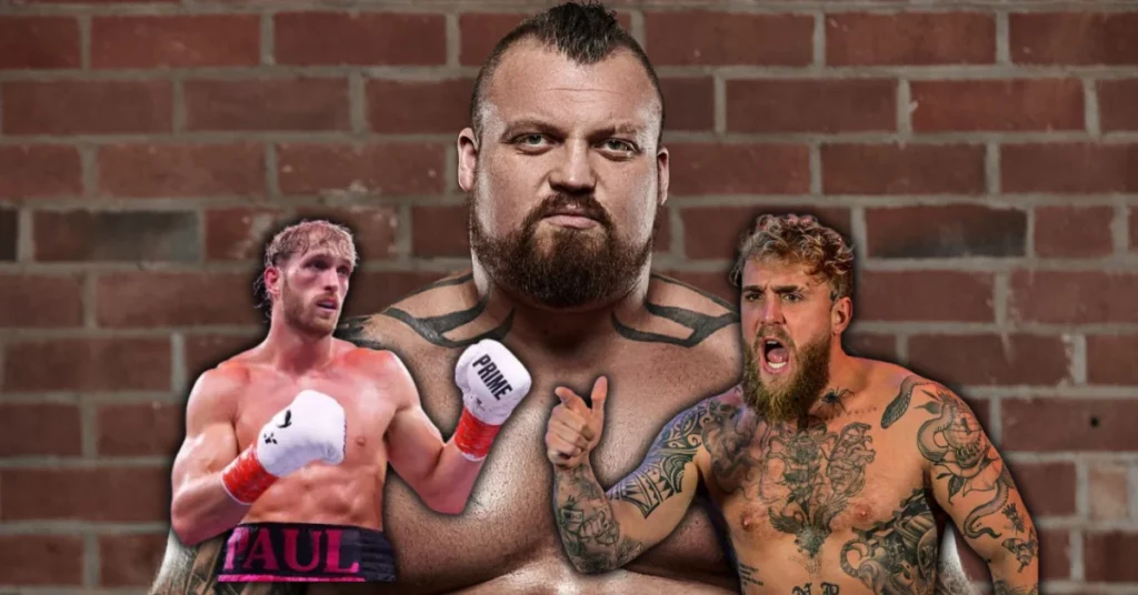 Eddie Hall, forzudo, mira serio entre los boxeadores Logan Paul (guante Prime) y Jake Paul, ambos con tatuajes. Pared de ladrillo.