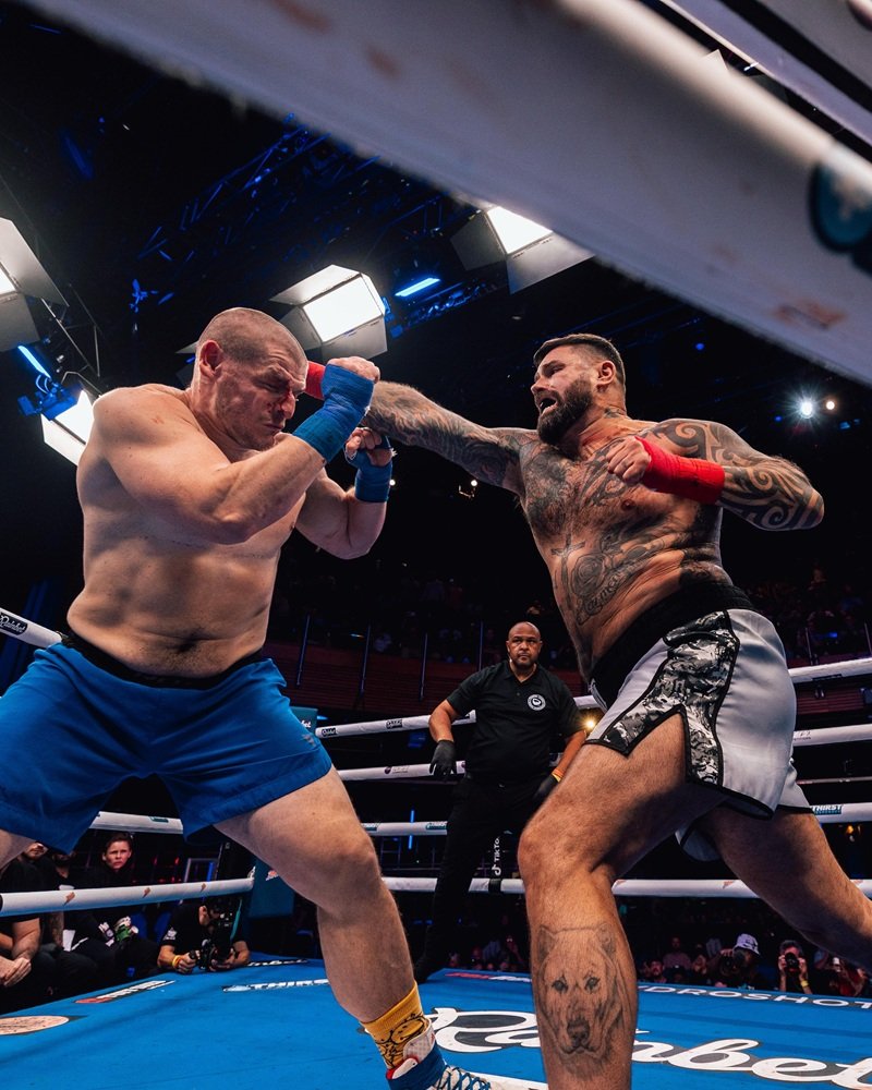Intenso combate de boxeo: púgil tatuado golpea con vendas rojas a su oponente con sangre en la cara, en un ring azul.