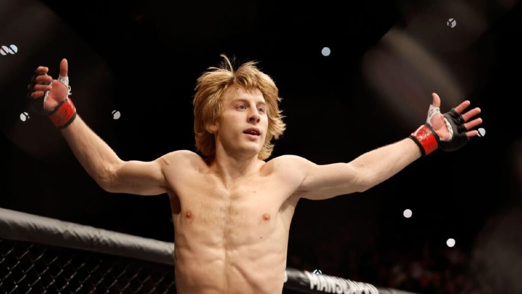 Luchador Paddy Pimblett rubio con guantes rojos celebra en el octágono, brazos abiertos y mirada hacia arriba.