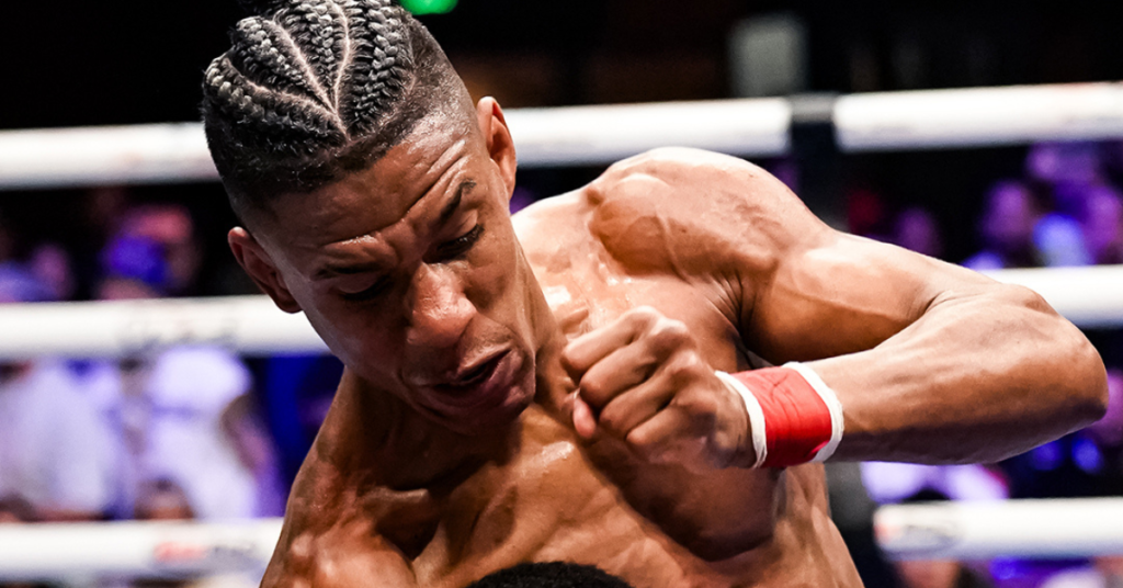 Boxeador con trenzas, músculos tensos y sudor. Rostro contraído por el esfuerzo o impacto en el ring, con vendaje rojo.