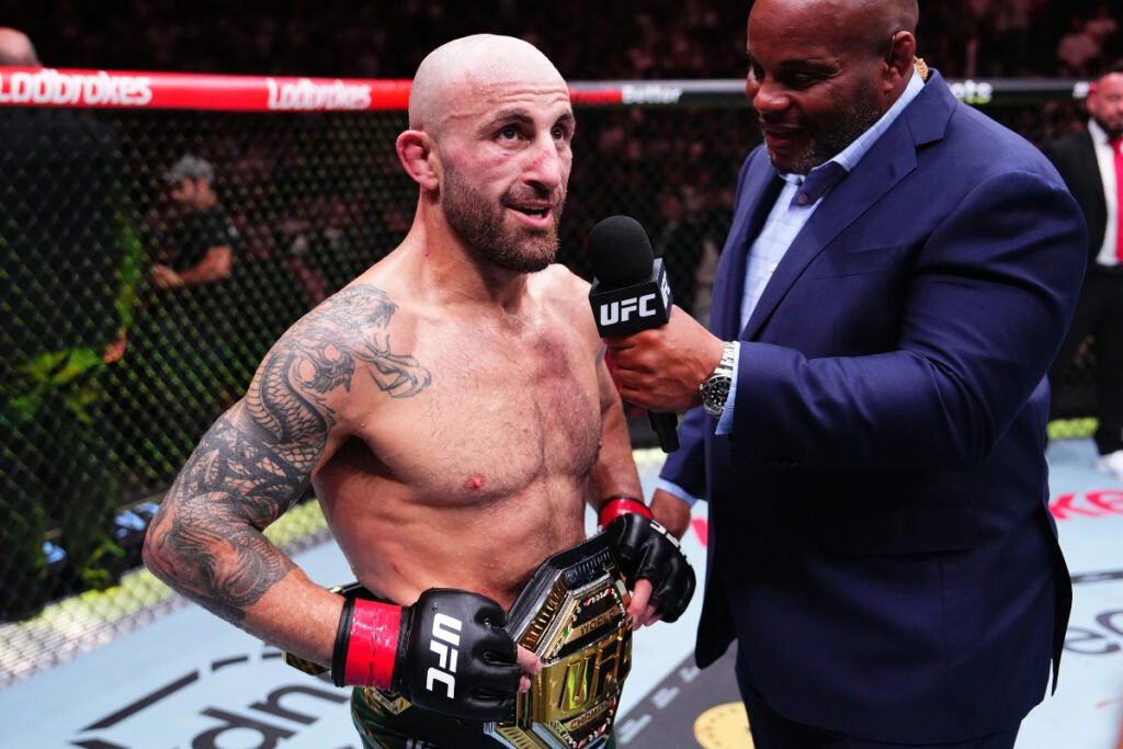 Alexander Volkanovski, campeón de UFC, con su cinturón y tatuajes, es entrevistado por Daniel Cormier con micrófono en el octágono.